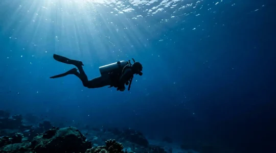 Plongeur en position horizontale parfaite à 20 mètres de profondeur, avec un manomètre visible, entouré de bulles dans une eau cristalline