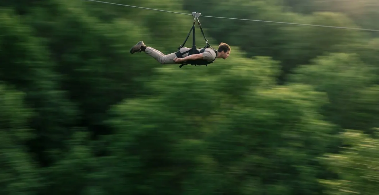 Personne en position aérodynamique compacte sur une tyrolienne, bras le long du corps, au-dessus d'une forêt floue